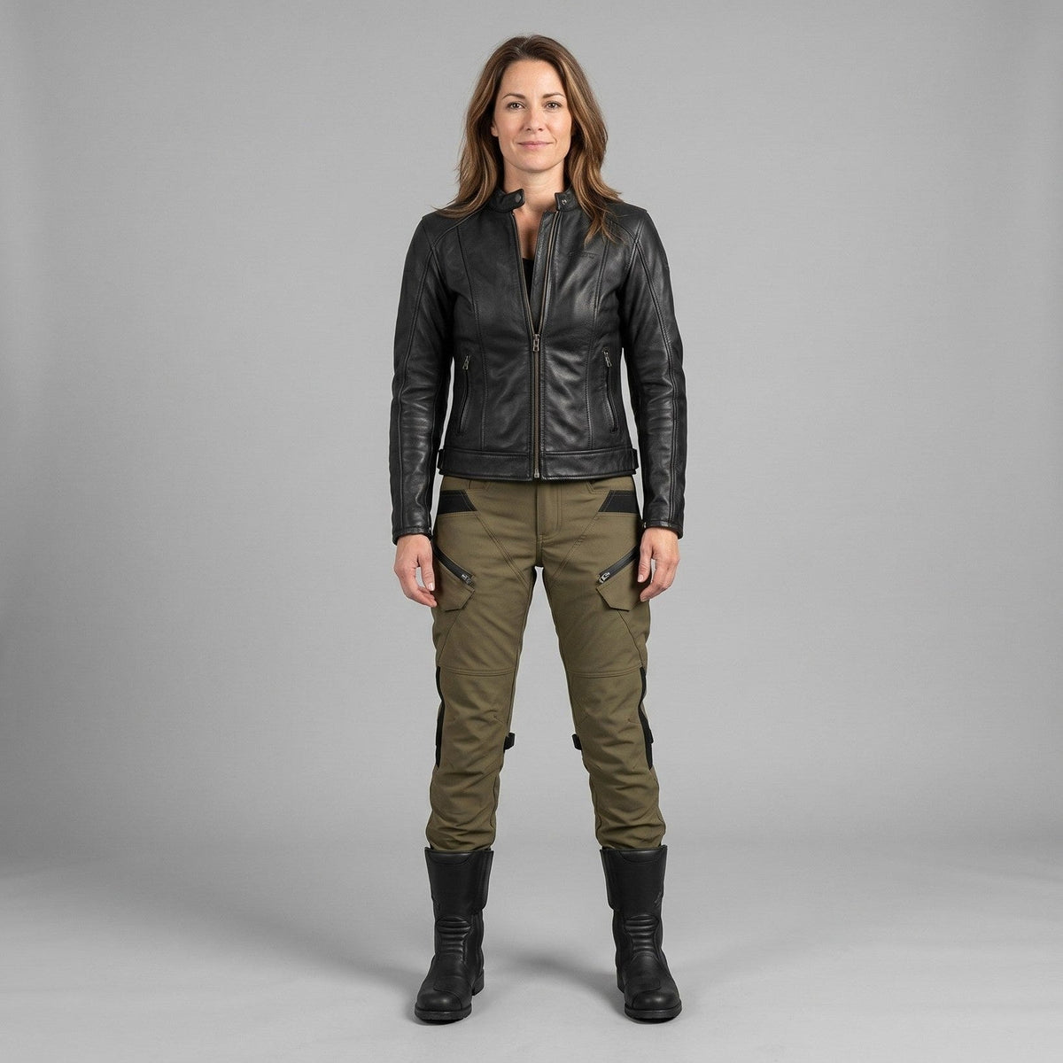 Woman standing in olive green Ride Unmarked RoadVault riding pants with a black leather jacket and tall black riding boots, studio shot on a grey background