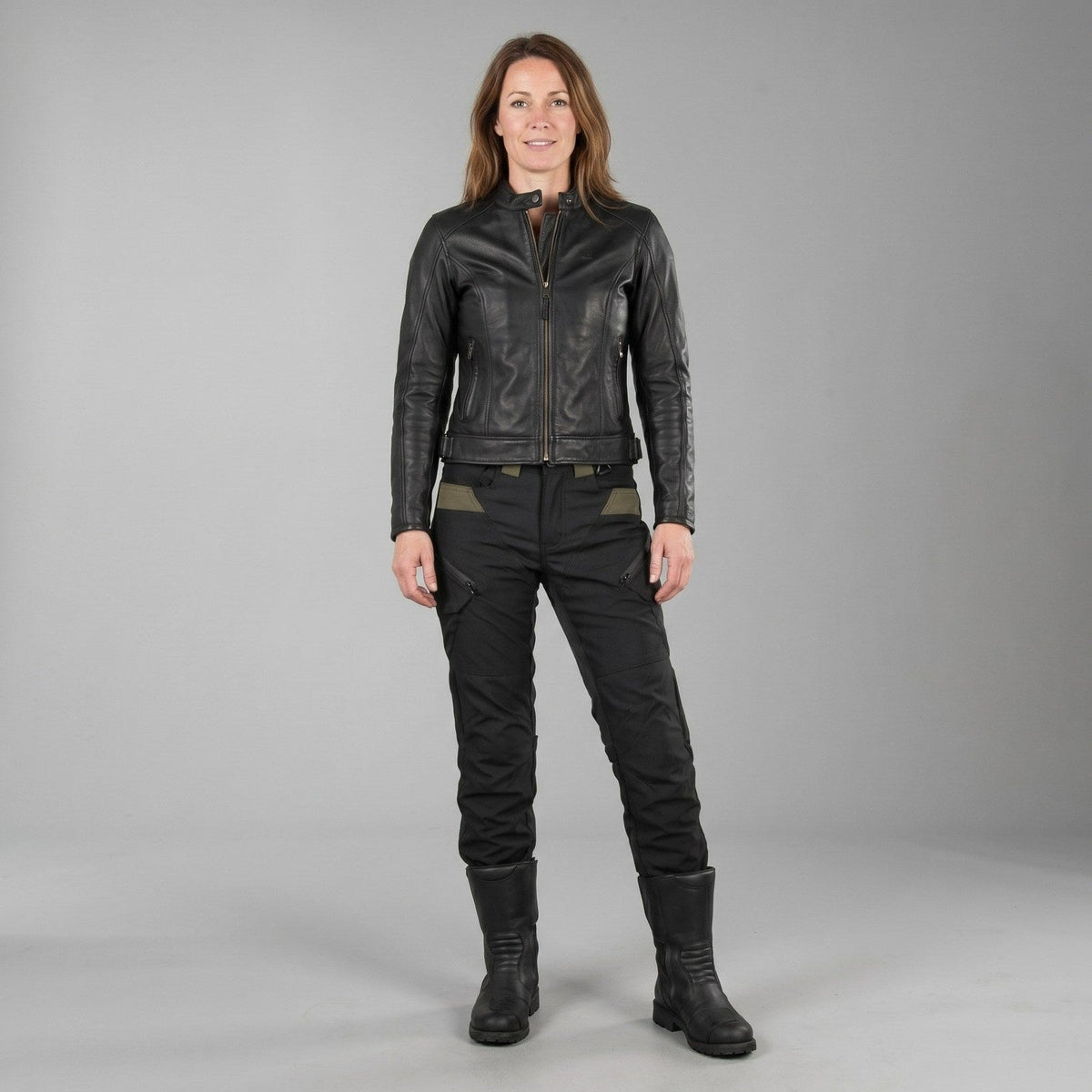 Woman in a black leather jacket and black cargo riding pants with olive panels at the waist, standing in tall black boots against a plain gray studio background
