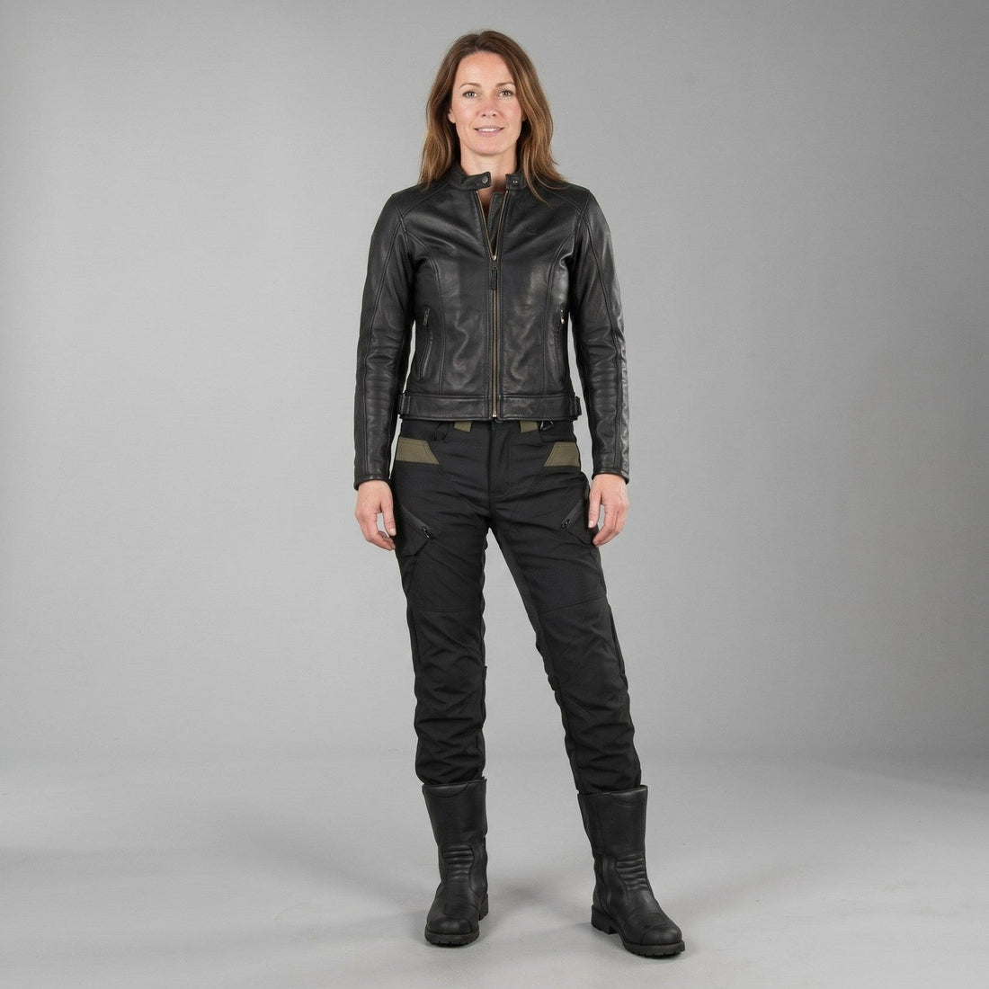 Woman in a black leather jacket and black riding pants with olive hip panels, the legs tucked into tall black boots, standing against a plain gray studio background