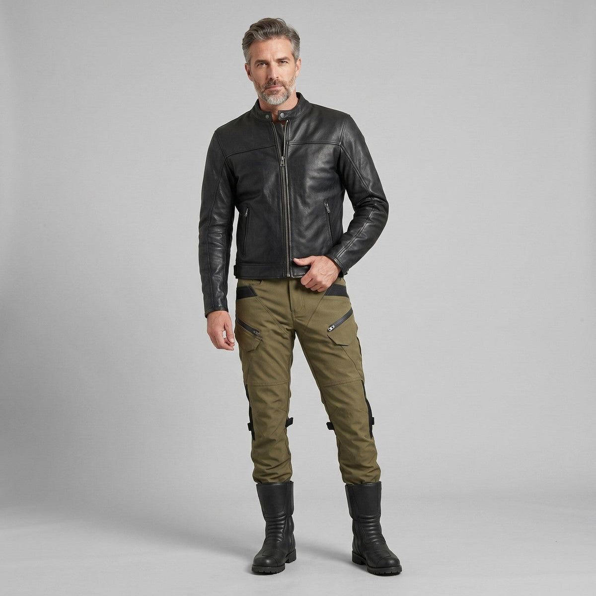Man in a black leather jacket, army green cargo-style riding pants and tall black boots against a plain gray studio background