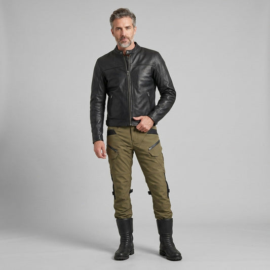 Man in a black leather jacket, army green cargo-style riding pants and tall black boots against a plain gray studio background