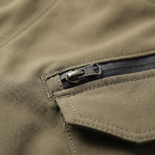 Close-up of olive riding pants with a stitched cargo pocket flap and an antique-brass zip pull on the zipped pocket above it